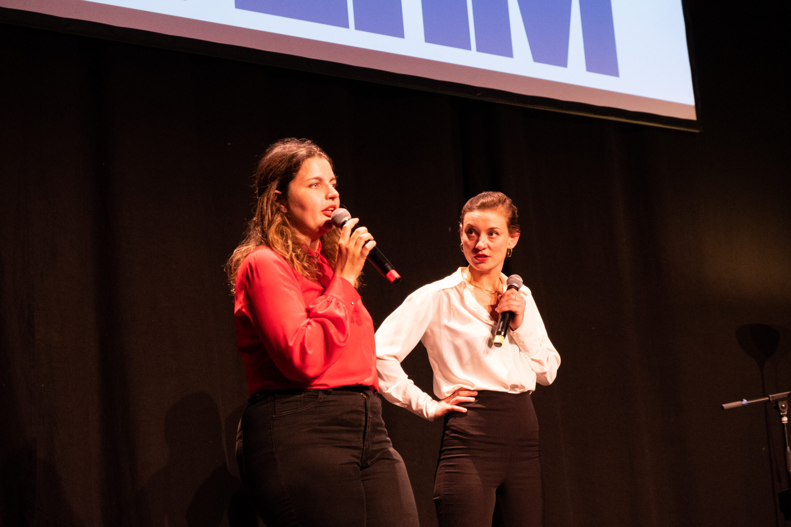 Zwei Personen stehen auf einer neutral ausgeleuchteten Bühne, einander zugewandt. Beide halten Mikros in der Hand. Eine trägt ein rotes Oberteil, die andere ein weißes Hemd. Beide dunkle Hosen.