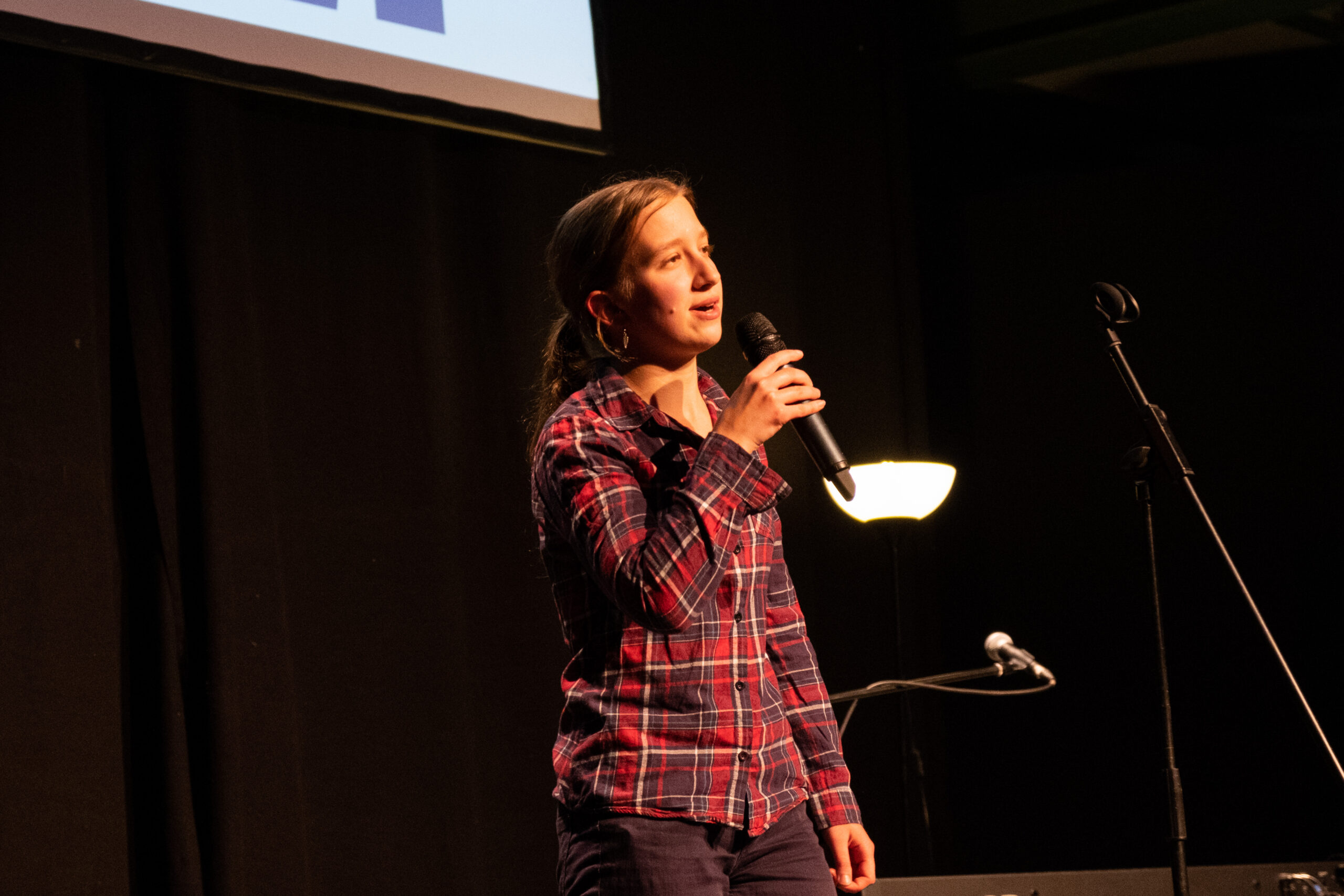 Eine Person in einem rot-karierten Hemd steht auf einer neutral ausgeleuchteten Bühne und spricht zu einem nicht sichtbaren Publikum.