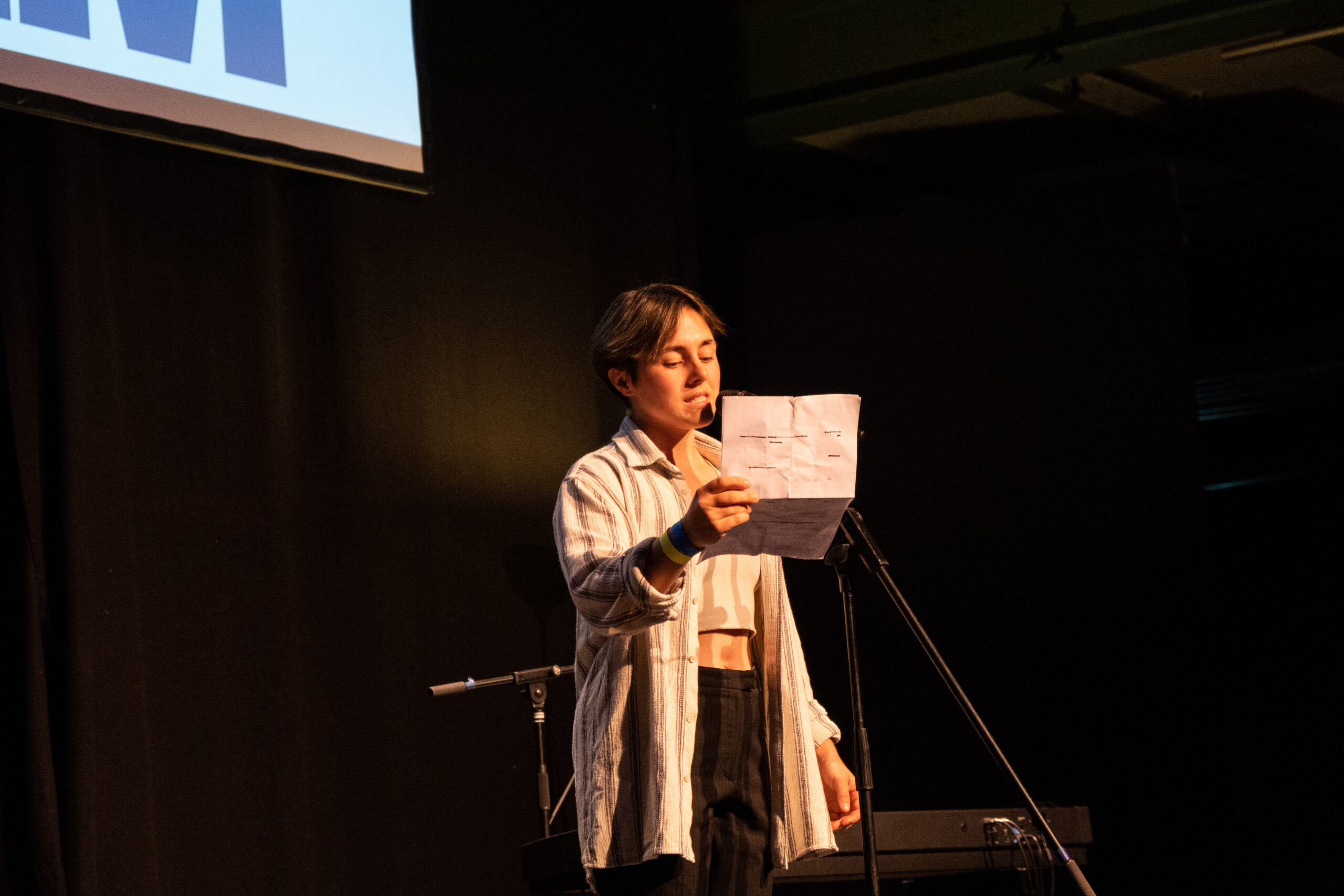 Eine Person in einem kurzen Oberteil, einem hellen Cardigan darüber und dunkler Hose steht vor einem Mikrofon und hält ein Papier in der Hand.