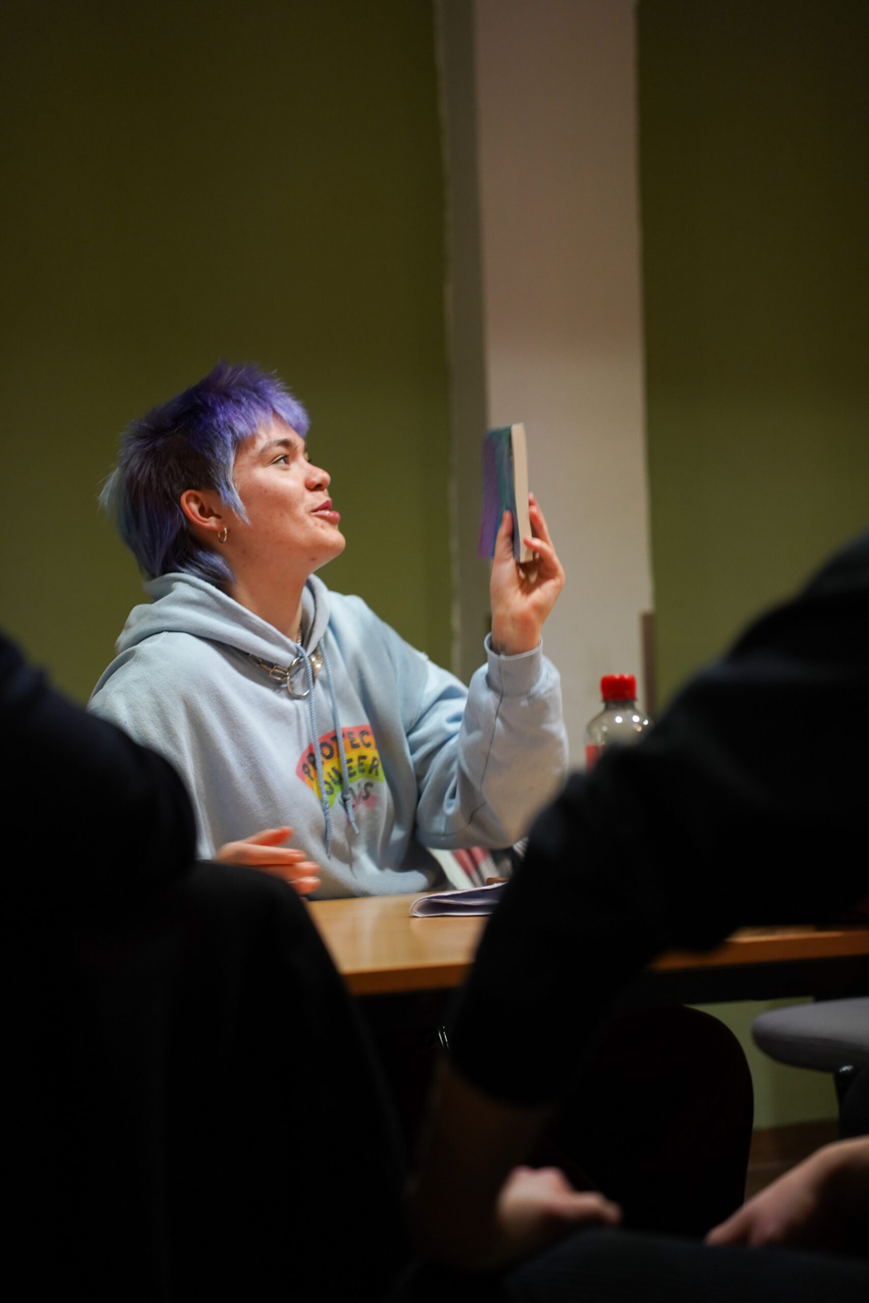 Eine Person mit hellen, lila Haaren hat ein dünnes Buch in der Hand und starrrt mit einem sehr übertriebenen Gesichtsausdruck darauf. Sie trägt einen grauen Hoodie und sitzt an einem Tisch.