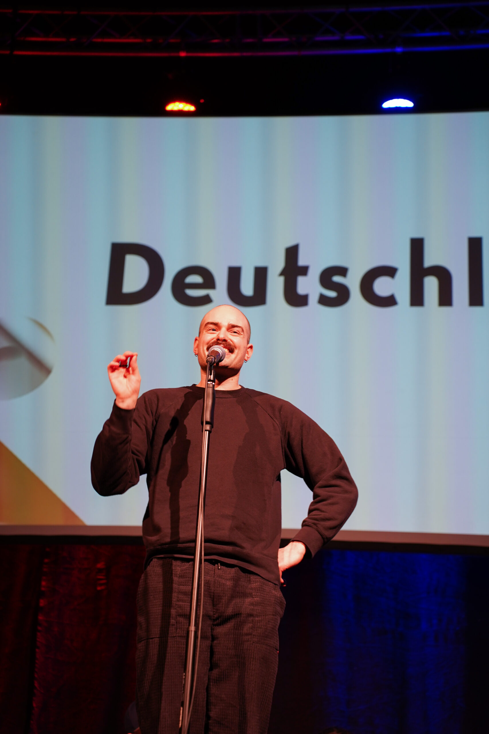 Eine glatzköpfige Person mit Schnauzbart steht auf einer Bühne vor einem Mikrofon. Sie trägt einen braunen Zweiteiler, der an einen Jogginganzug erinnert.