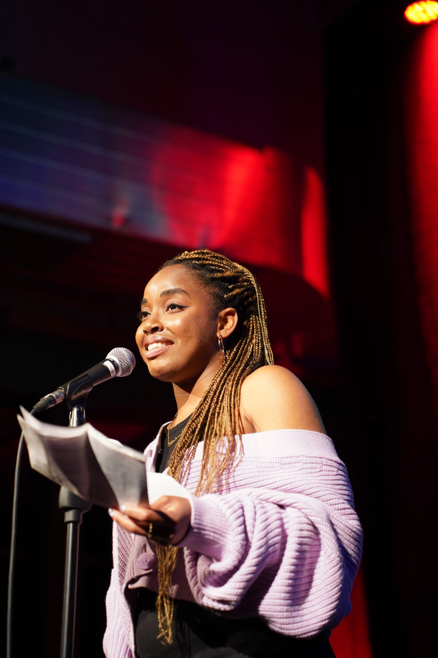 Eine Schwarze Person mit langen, geflochtenen Braids steht vor einem Mikro. In der Hand einige Blätter Papier. Sie trägt einen flauschigen, rosa Pulli. Die Bühne ist erleuchtet.