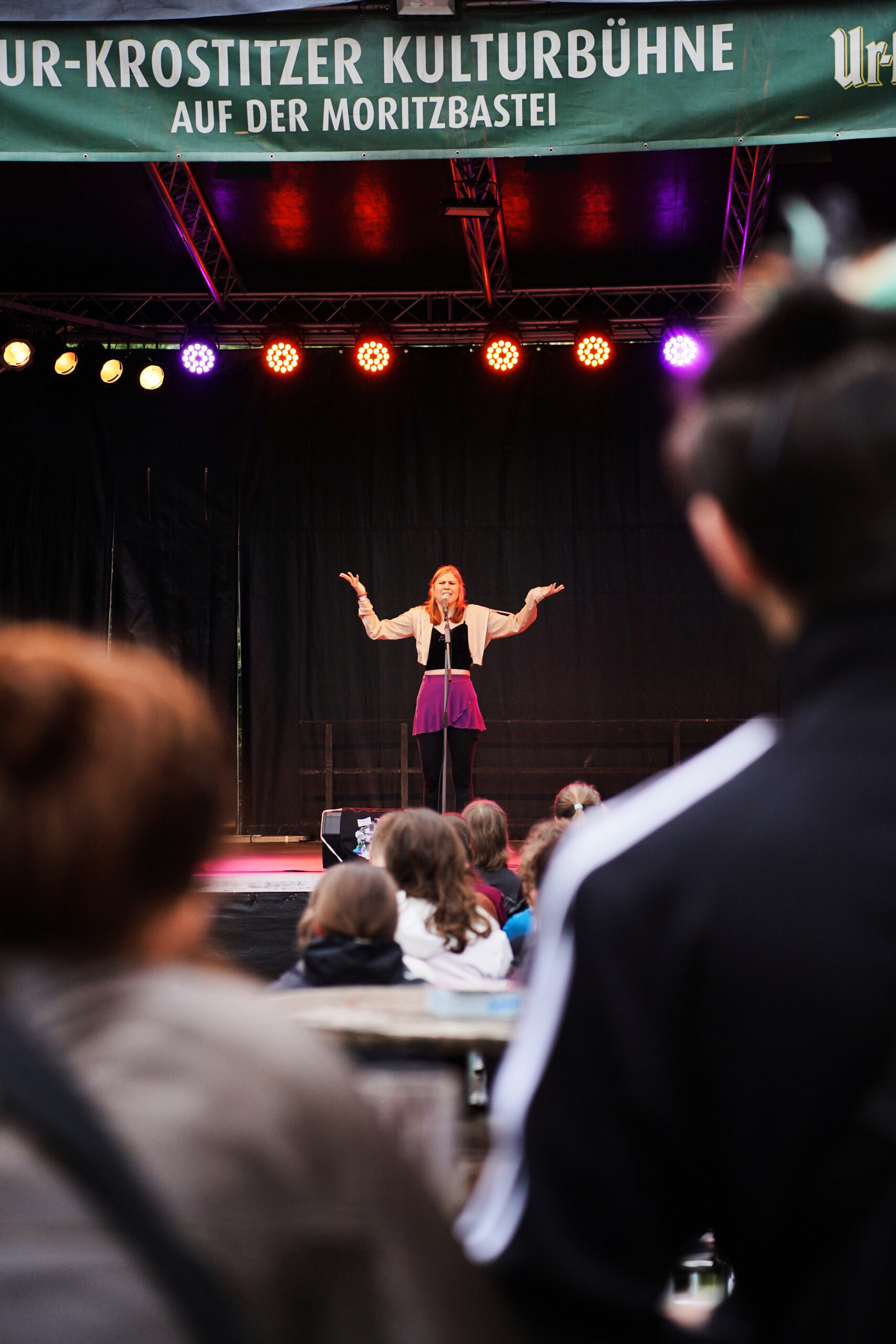 Eine Aufnahme aus dem Publikum heraus, bei der eine Person mit erhobenen Armen auf der Bühne steht. Sie trägt einen lila Rock und eine helle Jacke. Über der Bühne hängt ein grünes Banner, hier ist "Moritzbastei" gut zu erkennen.