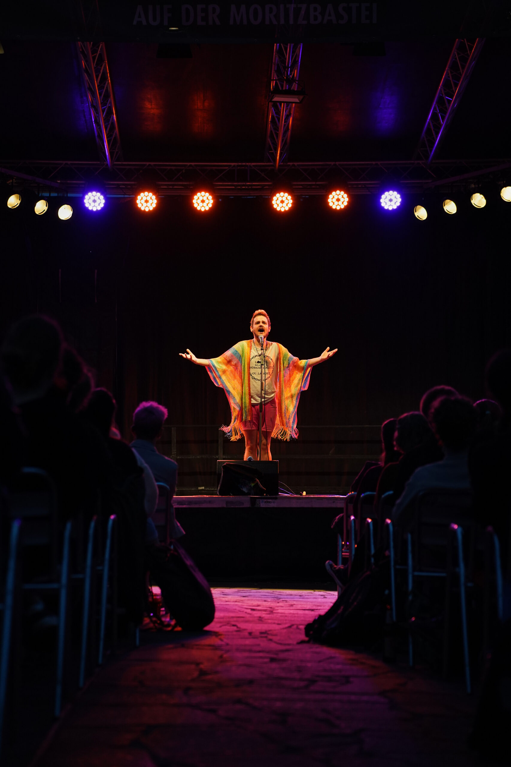 Eine dunkle Bühne, auf der eine Person in einem orangen Umhang mit erhobenen Armen zum Publikum spricht.