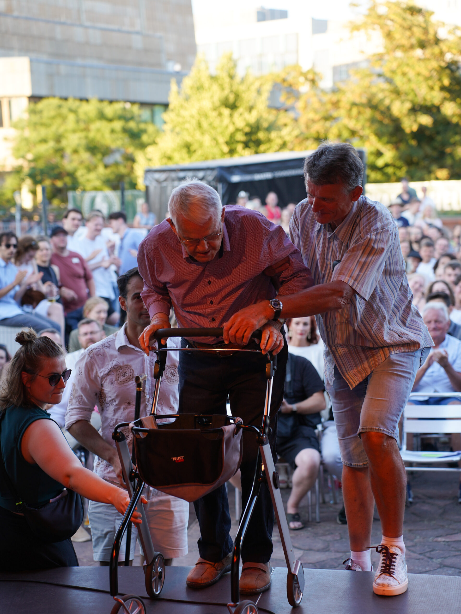 Eine ältere Person mit grauen Haaren hilft einer alten Person mit einem Rollator auf eine Bühne.