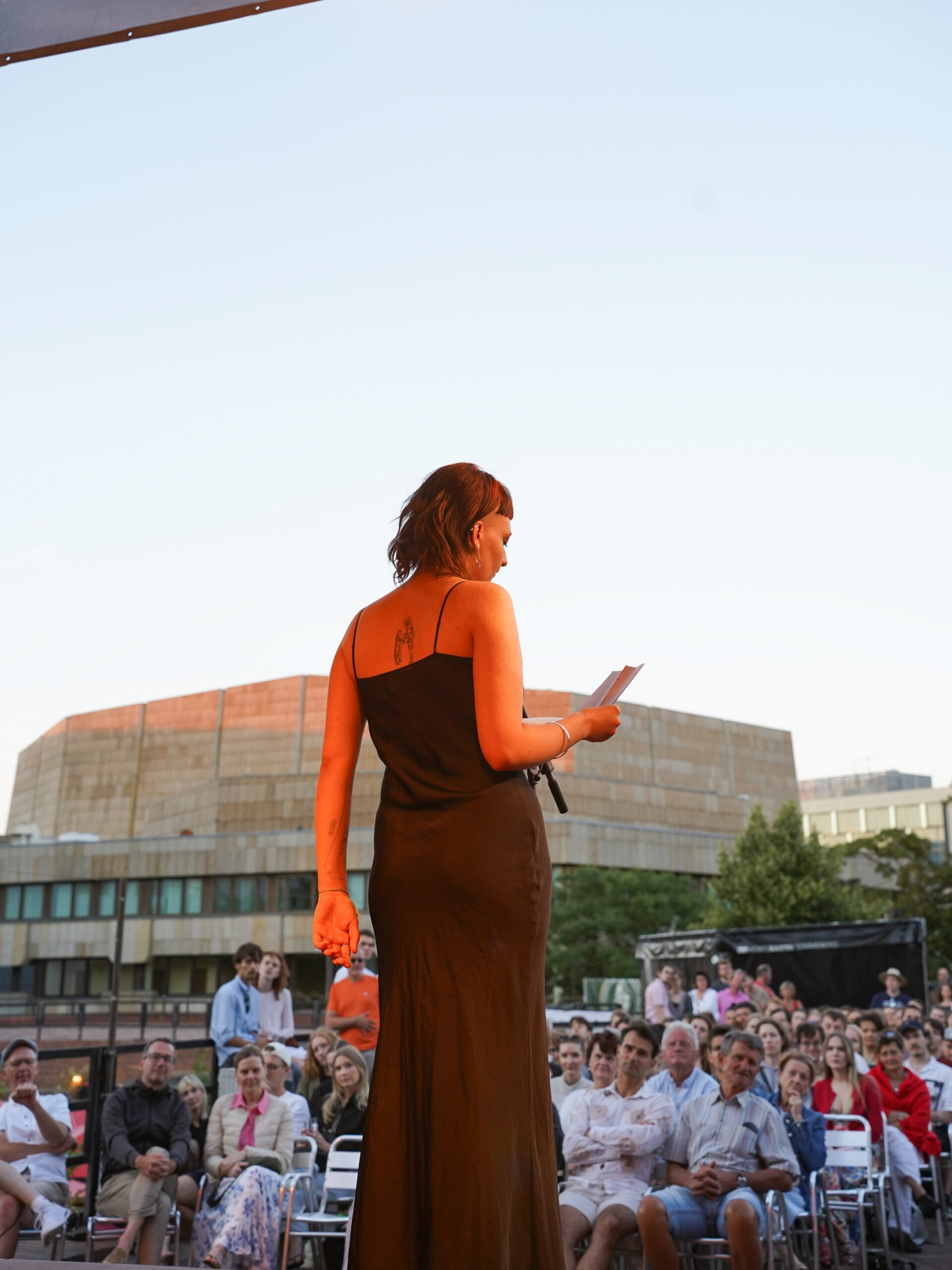 Eine Person in einem dunkelbraunen Kleid steht im Abendlicht auf einer Bühne. Sie hat etwas in der Hand, von dem sie offenbar abliest.